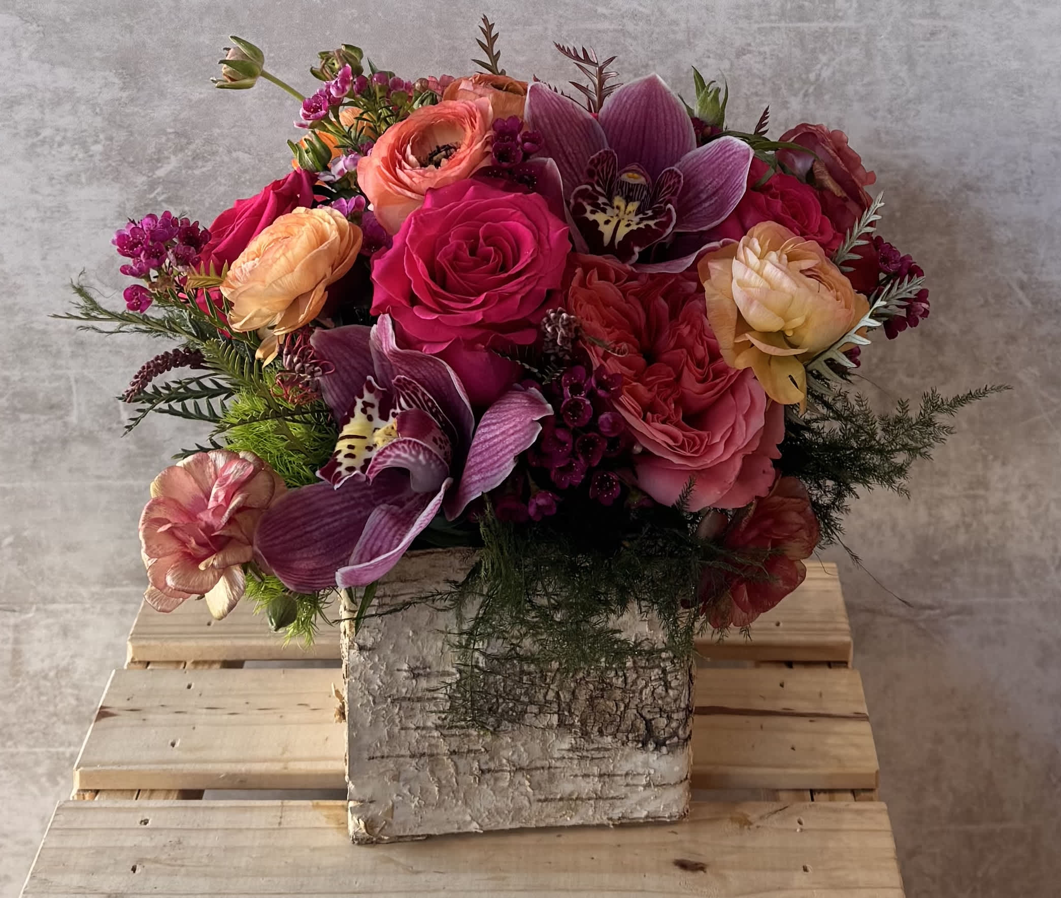 Bouquet of colorful flowers in a rustic vase on a wooden pallet against a gray wall.